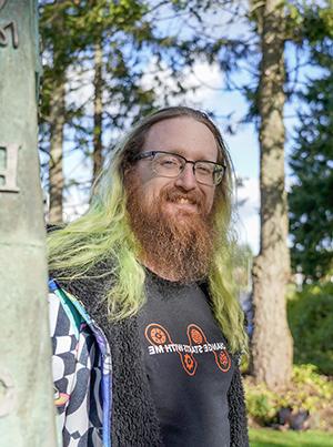 A man with green hair and a beard by the TCC peace pole smiling. 